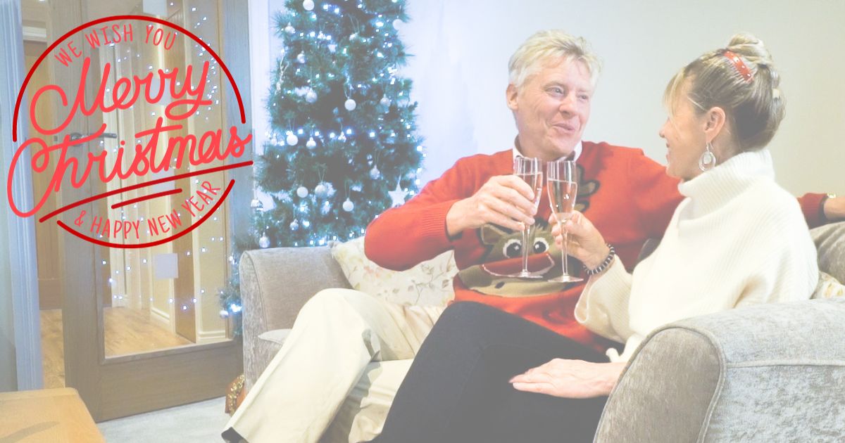 Couple sat on sofa cheers champagne glasses with Christmas tree in background and Merry Christmas greeting overlay